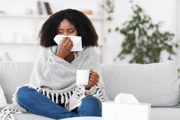 Sick afro woman wrapped in blanket blowing her running nose Fever, Cold, Flu Symptoms And Folk Medicine. Sick ill black woman covered in blanket blowing her running nose in a tissue sitting on sofa, drinking hot tea, holding cup, copy space. Taking medication tea hot drink photos stock pictures, royalty-free photos & images