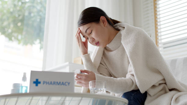 Sick asian woman sit under blanket acute severe headache on sofa at home. Female hold or check drug medicine capsule at home, suffer from flu. Cold or fever side effect after Covid19 vaccine shot. stock photo