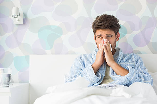 Sick man blowing his nose while sitting on bed stock photo