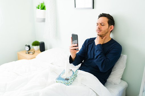 Latin man in bed talking to a virtual doctor during an online video call on his smartphone because of a sore throat