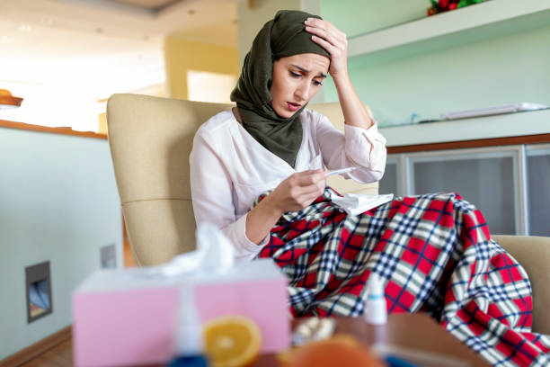 Sick Woman with Flu Checking the Temperature A sick young woman of Arabian ethnicity is sitting on the sofa at home in a bad health condition. A beautiful Muslim woman is checking the temperature of her body with a thermometer. hot middle eastern women stock pictures, royalty-free photos & images