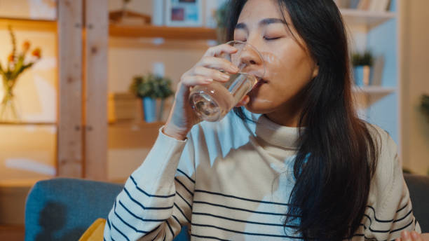 薬を飲む水のピルグラスを持っている病気の若いアジアの女性は、自宅の夜にソファに座っています。 - 夜　水　飲む ストックフォトと画像