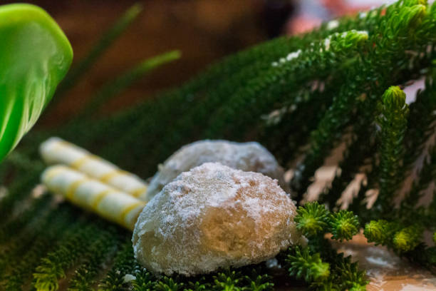 Side angle closeup photo of a one Snow Ball Christmas Cookie placed on a natural green leave with two sweet sticks and a partly visible green plastic bowl in the blurred background stock photo