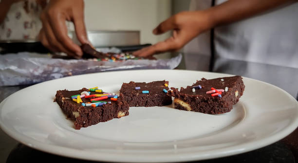 Side angle photo of three(3) pieces of Chocolate Biscuit Sprinkle Cakes arranged on a white plate. Woman's hands are visible in the background preparing & holding a piece about to place on the plate stock photo
