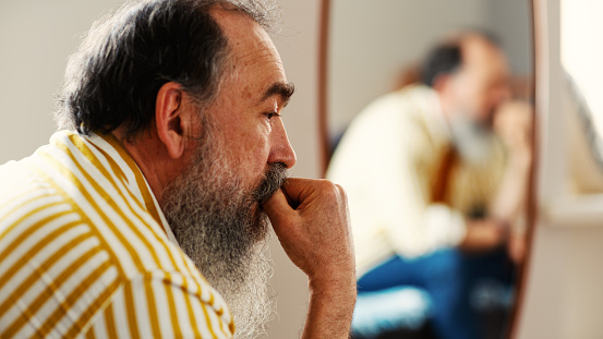 Side view head and shoulders shot of depressed pensive senior man with gray beard sitting with hand on chin near mirror at home or in hotel, feeling lonely and thinking what to do