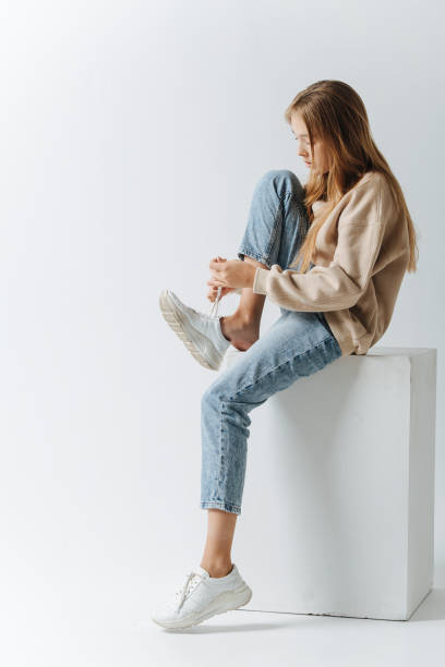 Side view of a teenage girl lacing her sneakers. Sitting on a white cube stock photo