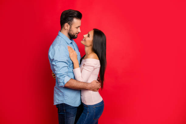 Side view of caucasian, attractive, smiling couple - bearded man embrace his charming, cute, pretty woman, looking to each other while standing over red background, 14 february Side view of caucasian, attractive, smiling couple - bearded man embrace his charming, cute, pretty woman, looking to each other while standing over red background, 14 february falling in love stock pictures, royalty-free photos & images