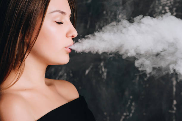 Side view of cute trendy girl exhaling big clouds of smoke from e-cigarette, standing over dark background, indoors.  little girl smoking cigarette stock pictures, royalty-free photos & images