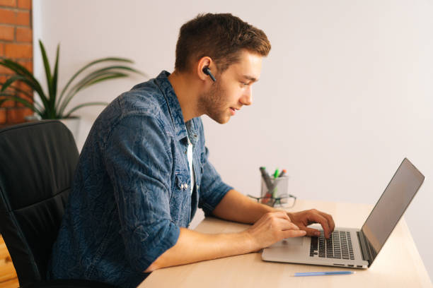 Side view of handsome young student in wireless earphone with microphone communicating via video call using laptop. stock photo