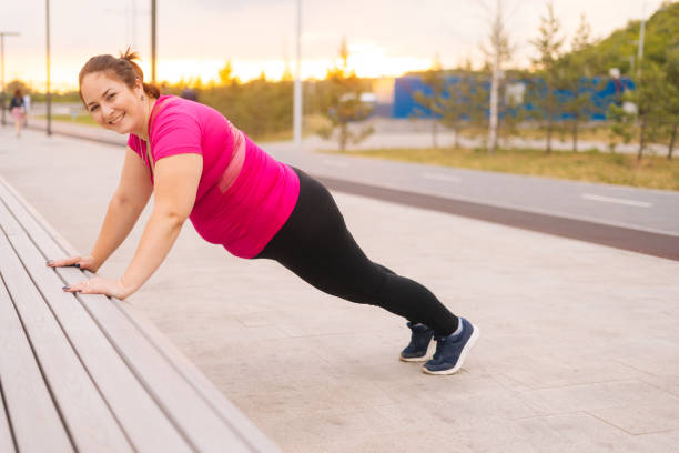 Side view of smilingfat young woman training doing push ups on street bench in city park at summer morning, looking at camera. stock photo