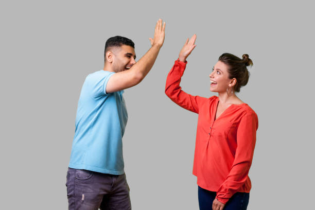 Side view portrait of excited amazed young couple in casual wear giving high five. isolated on gray background Side view portrait of excited amazed young couple in casual wear standing saying hello and giving high five, friends greeting each other, glad to meet. isolated on gray background, indoor studio shot high five stock pictures, royalty-free photos & images