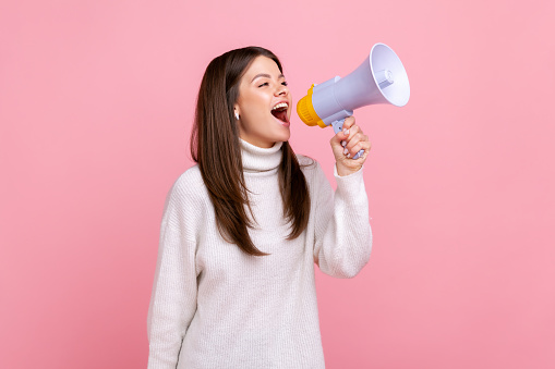 Side view portrait of pretty positive girl screaming in megaphone, announcing important information, wearing white casual style sweater. Indoor studio shot isolated on pink background.