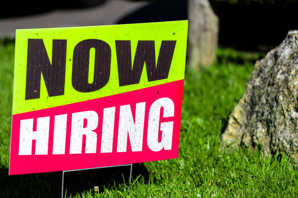 Sign text closeup for help wanted now hiring in English with red and green colors as signpost on grass for store shop business during corona virus covid 19 pandemic stock photo