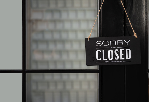 Signboard of tell sorry we are closed business and please come back again when situation improved with hanging to door front at shop.