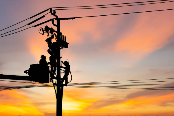 Silhouette of Electrician officer climbs a pole and uses a cable car to maintain a high voltage line system, Shadow of Electrician lineman repairman worker at climbing work on electric post power pole stock photo