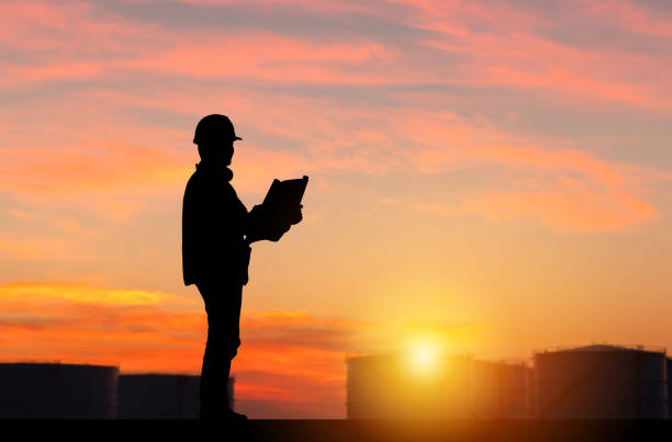Silhouette of Engineer under inspection and checking oil storage tank, Engineer man in waistcoats and hardhats and with documents in oil storage plant stock photo