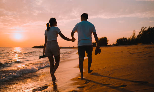 Silhouette of happy Asian couple hands holding and walking together on the beach while golden sunset time evening of summer vacation. stock photo