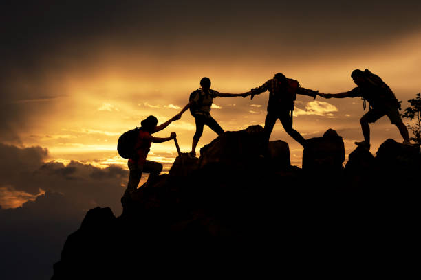 Silhouette of Hikers climbing up mountain cliff. Climbing group helping each other while climbing up in sunset. Concept of help and teamwork, Limits of life and Hiking success full. stock photo