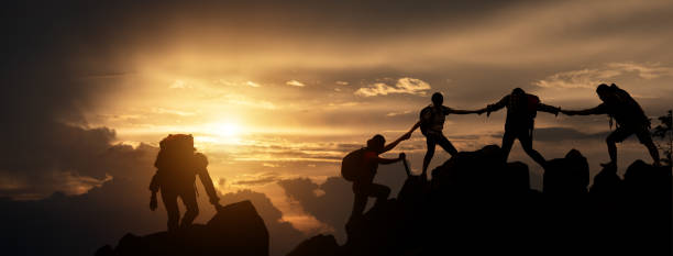 Silhouette of Hikers climbing up mountain cliff. Climbing group helping each other while climbing up in sunset. Concept of help and teamwork, Limits of life and Hiking success full. stock photo