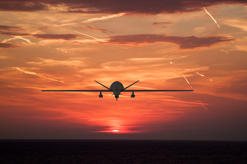 Silhouette of spy drone flying over the sea (UAV) and on background beautiful view of sun hiding behind sea surface.  sunset sky is orange with clouds and condensation traces