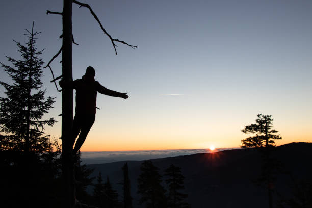 Silhouette of teenager dressed in winter clothes standing on branch and enjoying his feeling of victory from uprising of the mountain and looking at the final rays of the sun in Beskydy mountains stock photo