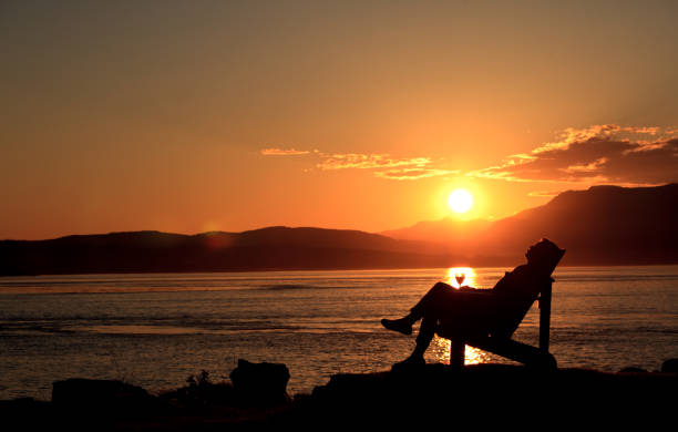 Silhouette of Woman Relaxing in Adirondack Chair Sipping Wine at Sunset by Ocean stock photo