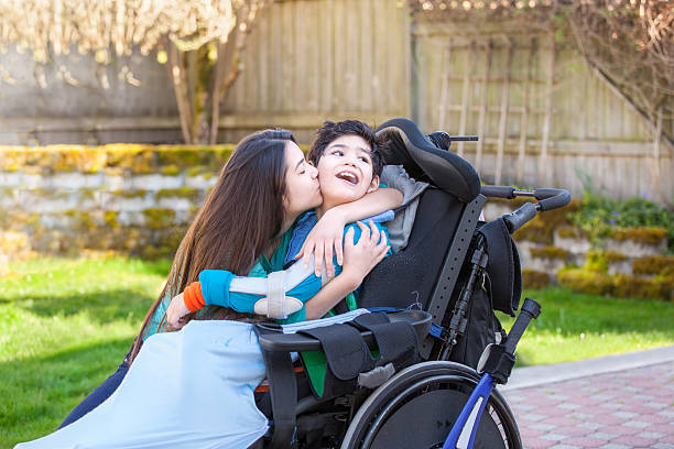 Sister kissing and hugging disabled little brother in wheelchair outdoors stock photo
