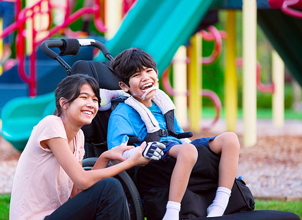 Sister sitting next to disabled brother in wheelchair at playground stock photo
