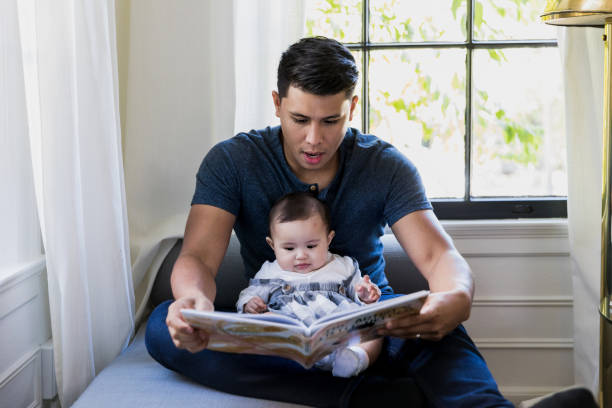 Sitting by window, dad reads book to baby As they sit by the window, the mid adult dad reads a book to his baby daughter. father and child reading stock pictures, royalty-free photos & images