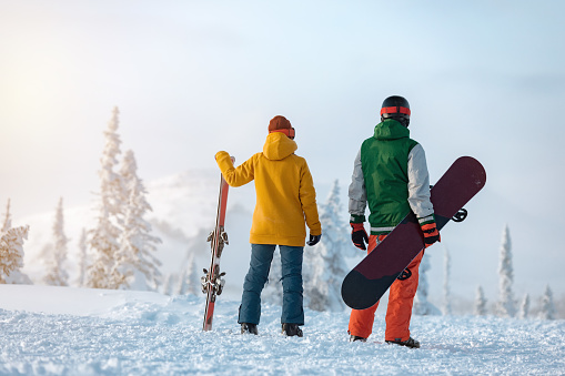 Female skier and male snowboarder are standing on background of snowcapped mountain at sunset time. Ski resort concept