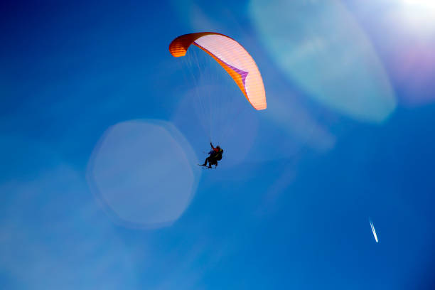 Skier Paragliding with skis using a selfie stick while flying through bright lens flair with blue sky and plane trails in the sky stock photo