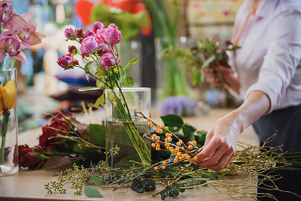 Skillful florist making bouquet in workshop Close up of female hand is taking branch of rowan to join bunch of flowers. Focus on roses and other plants on table in flower shop flower arrangement stock pictures, royalty-free photos & images