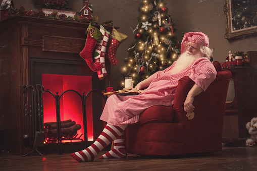 Santa Claus sits in a sofa chair in front of the fireplace in his living room, wearing pajamas with a plate of cookies and milk on his lap and a half eaten cookie in his hand, fast asleep, exhausted from delivering presents to all the boys and girls.