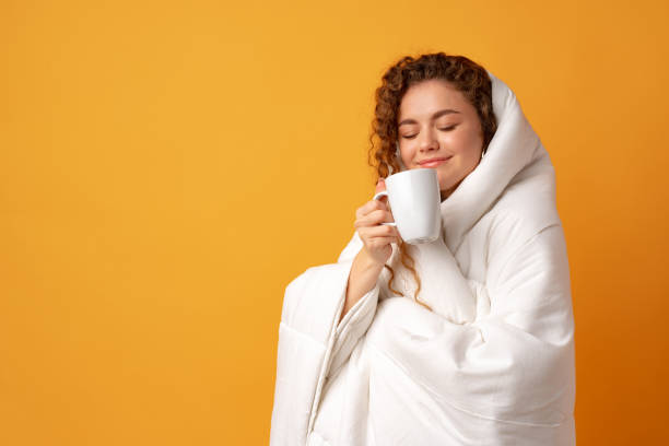 Sleepy young woman with curly hair covered in blanket and holding cup of coffee against yellow background stock photo