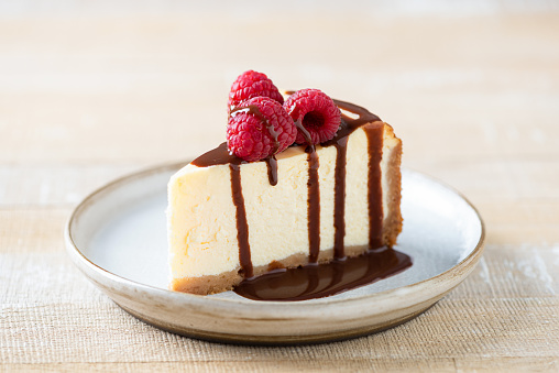 Slice of cheesecake with chocolate sauce and raspberries on a wooden table