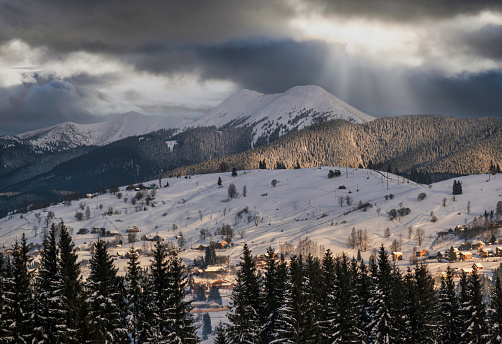 Small alpine village and winter snowy mountains in sunrise sun rays through clouds, Voronenko, Carpathian, Ukraine.