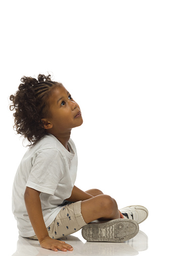 Small black boy in sneakers, shorts and shirt is sitting legs crossed on a floor and looking up. Side view. Full length studio shot isolated on white.