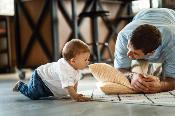 Small boy crawling while spending time with his father at home. Happy father talking to his small son who is crawling on the floor in the living room. crawling stock pictures, royalty-free photos & images