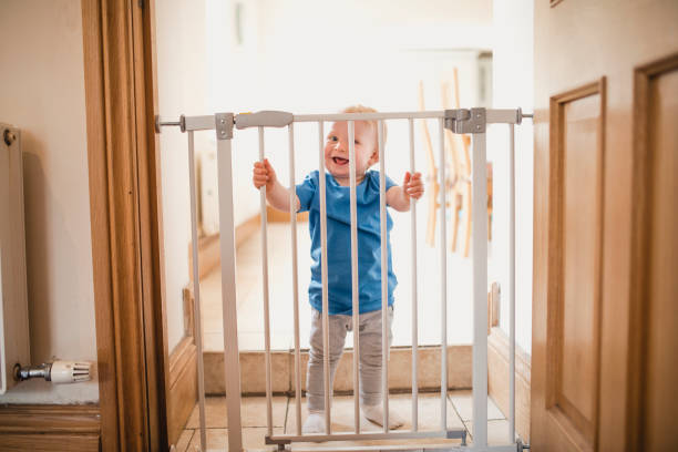 Small Boy Holds On To Safety Gate A joyful child smiles whilst holding on to a locked safety gate at home, he is wearing casual clothing and has a big grin on his face. gate photos stock pictures, royalty-free photos & images