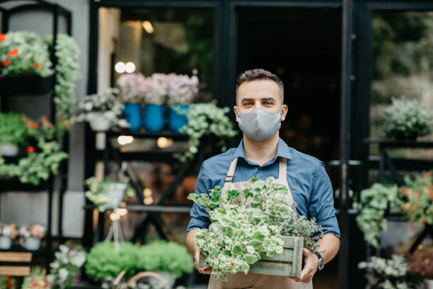 中小企業と営業日の開始。保護マスクを着用した男が外の植物の箱を取り出す - スモールビジネス ストックフォトと画像