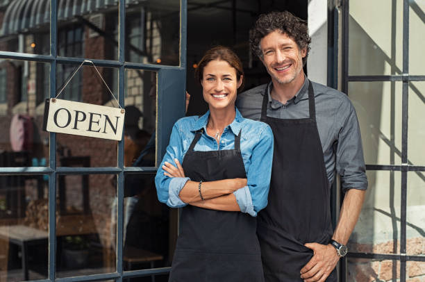 Small business owners couple Two cheerful small business owners smiling and looking at camera while standing at entrance door. Happy mature man and mid woman at entrance of newly opened restaurant with open sign board. Smiling couple welcoming customers to small business shop. small business stock pictures, royalty-free photos & images