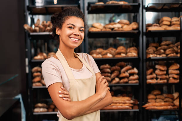 малый бизнес. молодая женщина в фартуке в пекарне магазин скрещенные руки позирует на камеру веселый - пекарь стоковые фото и изображения