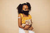 Small girl wearing protective face mask sitting with her teddy bear. Girl child with face mask on brown background after getting vaccination.