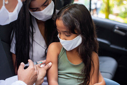 Close-up of a small girl wearing face mask receiving vaccination with mother in car. Family getting covid-19 vaccine at drive thru vaccination center in the city during pandemic.