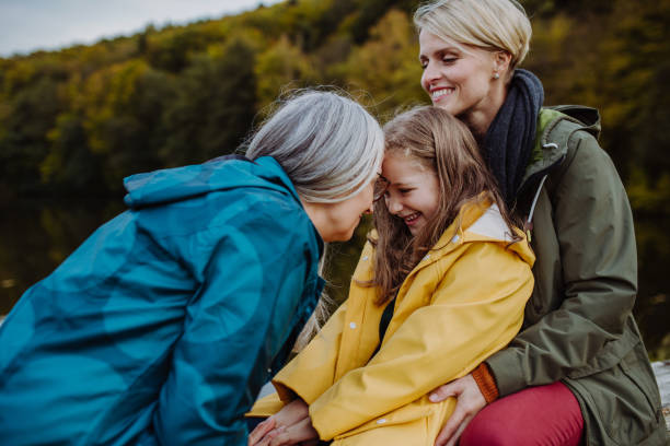 Small girl with mother and grandmother sitting on bench and having fun outoors by lake. stock photo