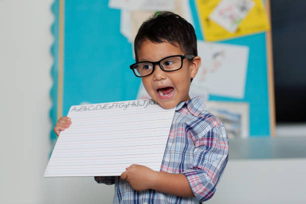 A smart boy wearing reading glasses and speaking or pronouncing words while holding up a blank writing board with a colorful bulletin board in the background. stock photo