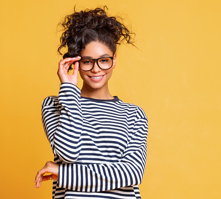 Clever ethnic female in striped garment smiling for camera and adjusting trendy glasses against yellow background