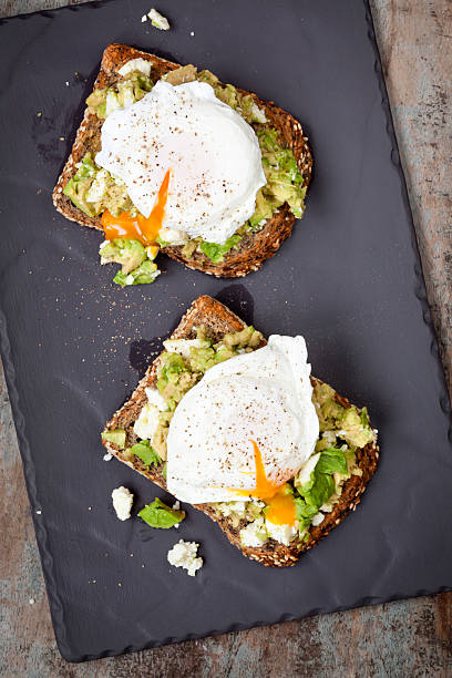 Smashed Avocado and Feta Toast with Poached Eggs Smashed avocado and feta cheese toast with poached eggs.  Overhead view, on dark slate. poached food stock pictures, royalty-free photos & images