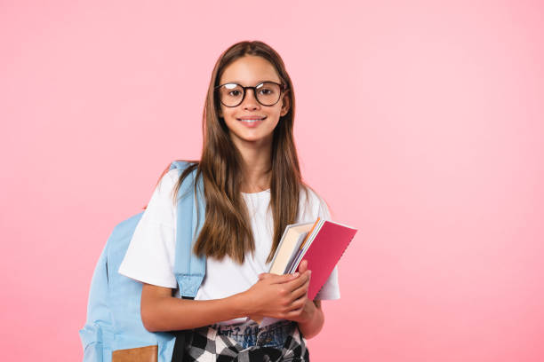 Smiling active excellent best student schoolgirl holding books and copybooks going to school wearing glasses and bag isolated in pink background stock photo