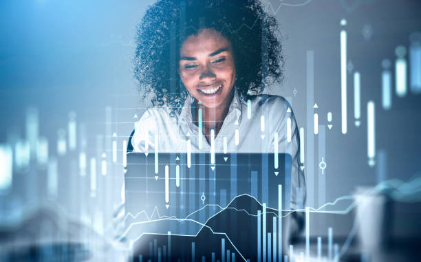 Smiling African American attractive young businesswoman in white shirt working on laptop. Forex graph hologram. Concept of internet trading and analytics. stock photo